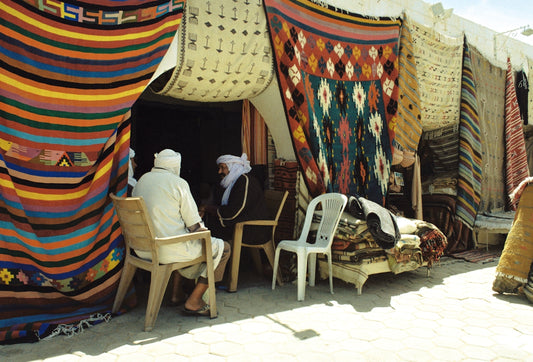 Men sit near colorful rugs displayed outside a shop